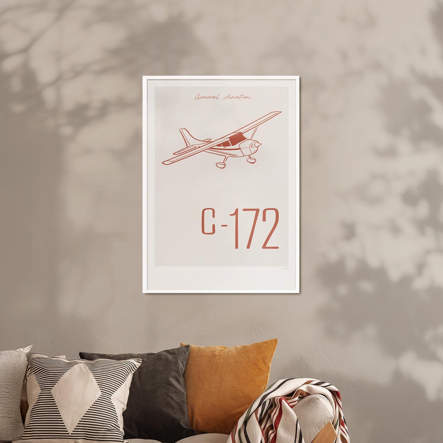 Framed poster hanging on a taupe wall above a sofa with multiple pillows. It shows a light aircraft illustration with the words General Aviation and C-172 in red
