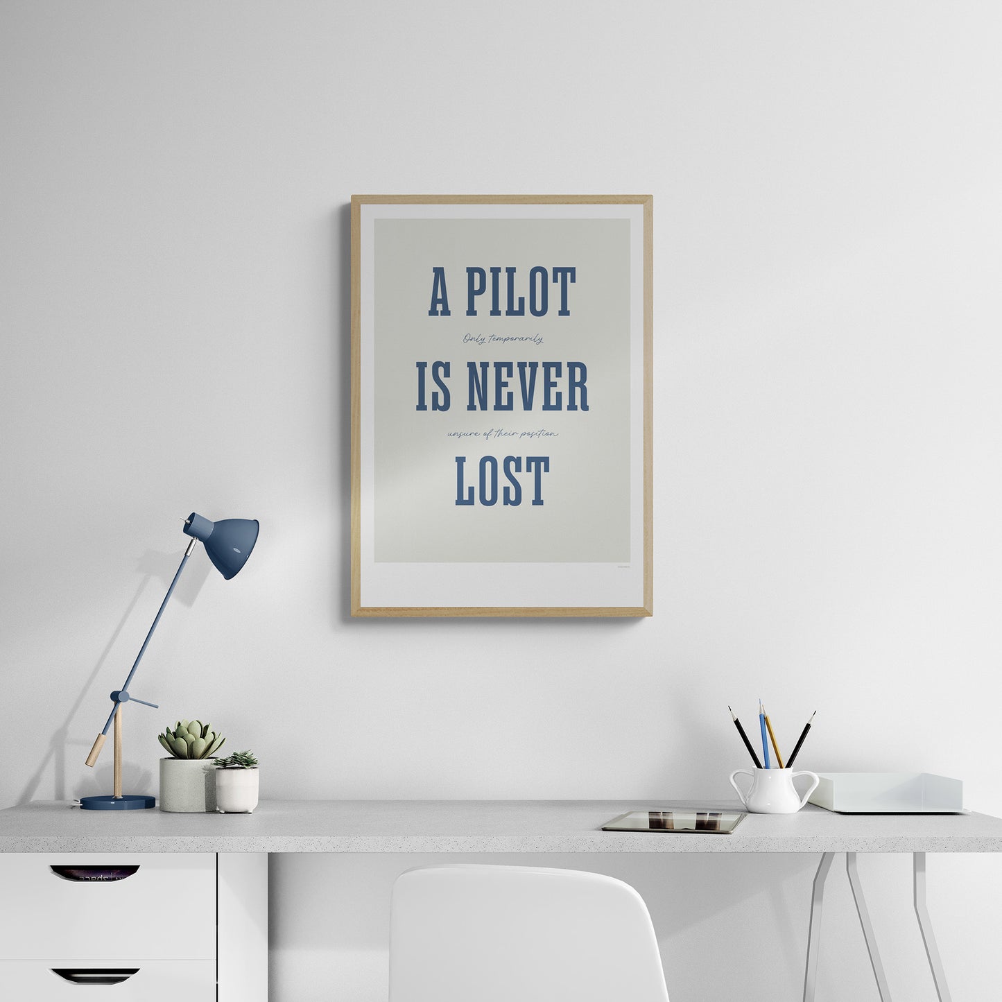 A framed print hanging in a home office over a white desk and chair. It shows the words - A Pilot Is Never Lost - in large blue letters and the words - Only temporarily unsure of their position - in in black handwritten style