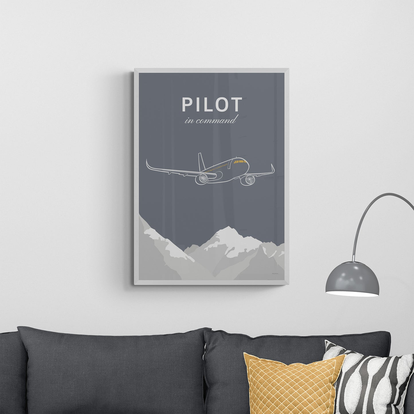 A framed print over a dark grey sofa with cushions next to a lamp. It shows a white illustration of a passenger aircraft over mountains on a grey background and the words pilot in command