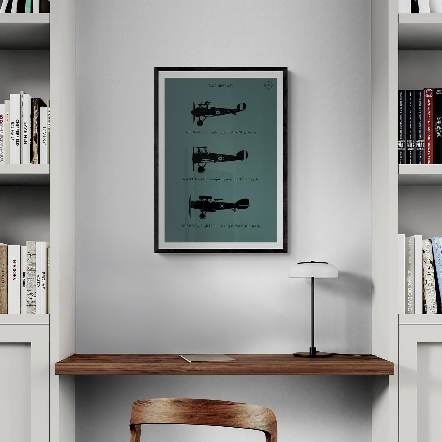 A framed print hanging in a home office over a wooden desk and chair next to 2 bookshelves. It shows the profiles of three WW1 period aeroplanes in a black silhouette style over a dark green background