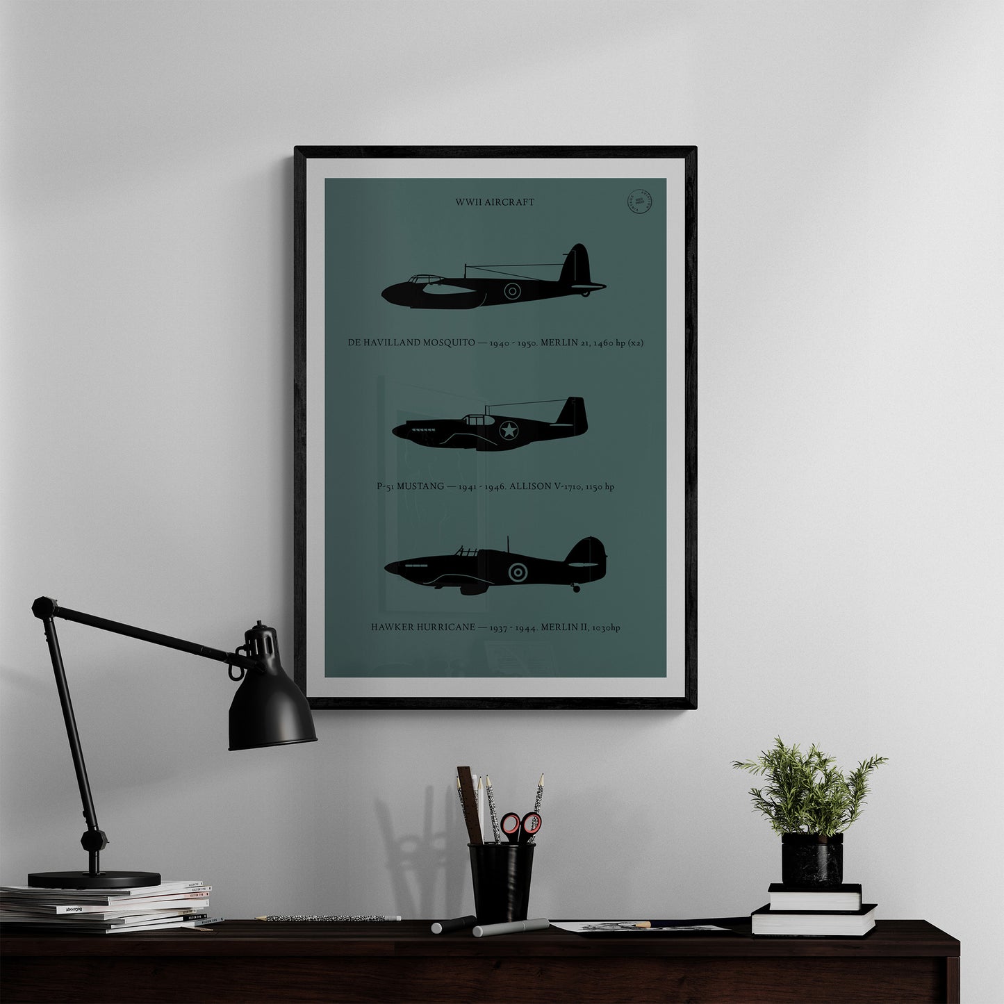 A framed poster hanging in a home office over a dark wood desk with books and a lamp. It shows the profiles of three WW1 period aeroplanes in a black silhouette style over a dark green background