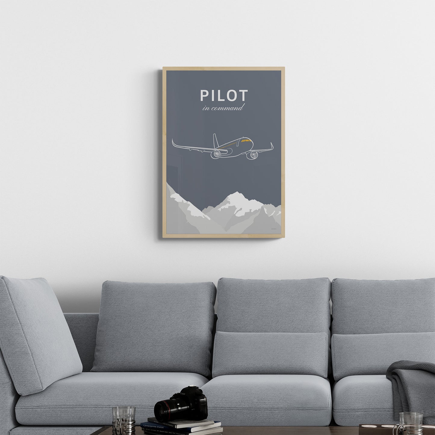 A framed print hanging in a living room over a grey sofa. It shows a white illustration of a passenger aircraft over mountains on a grey background and the words pilot in command