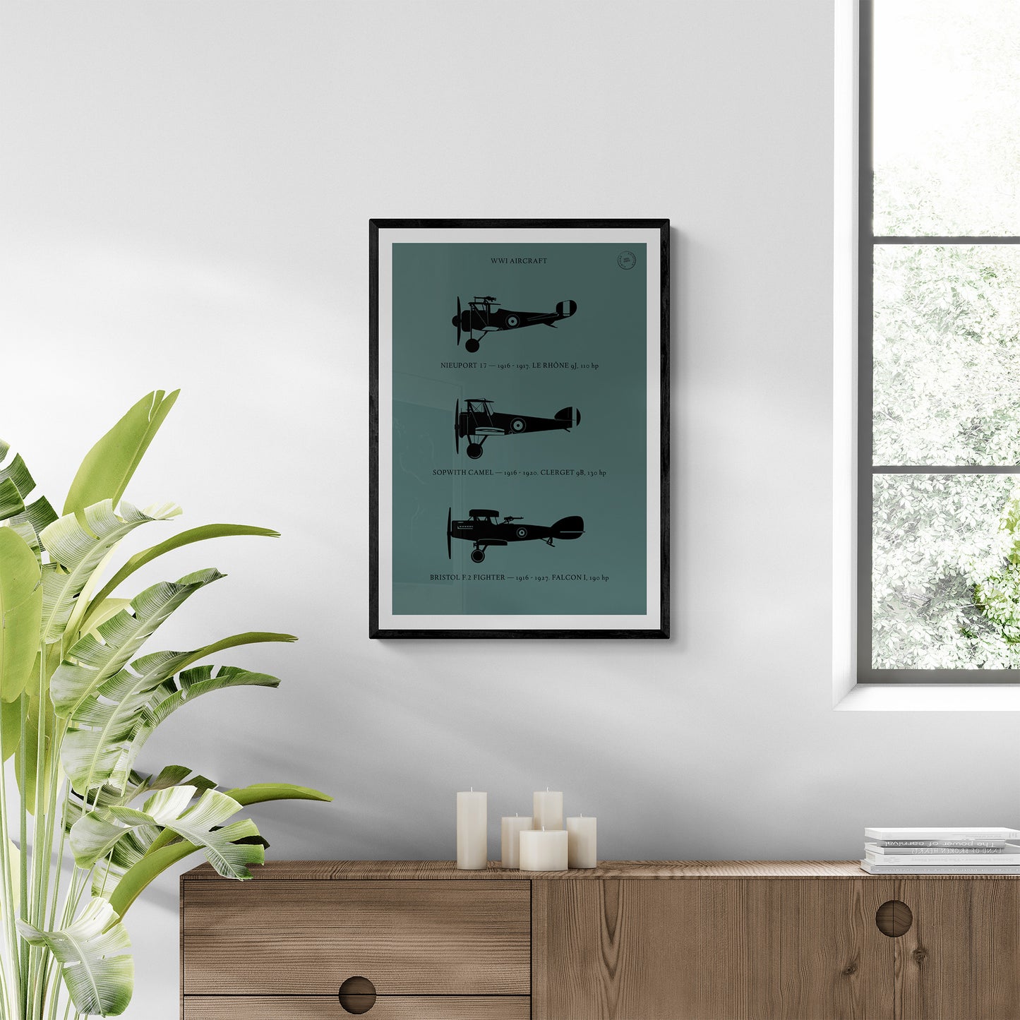 A framed print hanging in a modern brightly lit room over a wooden console table and next to a plant. It shows the profiles of three WW1 period aeroplanes in a black silhouette style over a dark green background