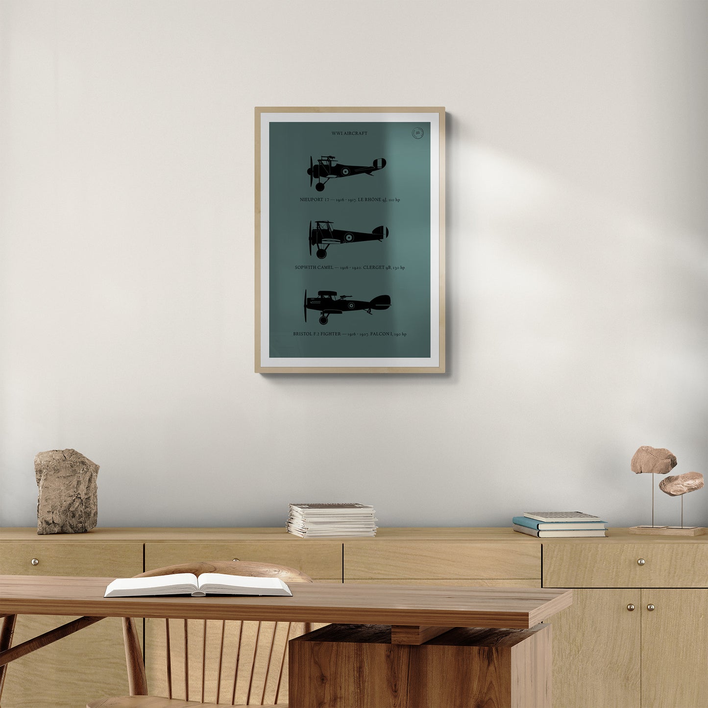 A framed print hanging in a home office over a wooden console table with books and a wooden desk and chair. It shows the profiles of three WW1 period aeroplanes in a black silhouette style over a dark green background