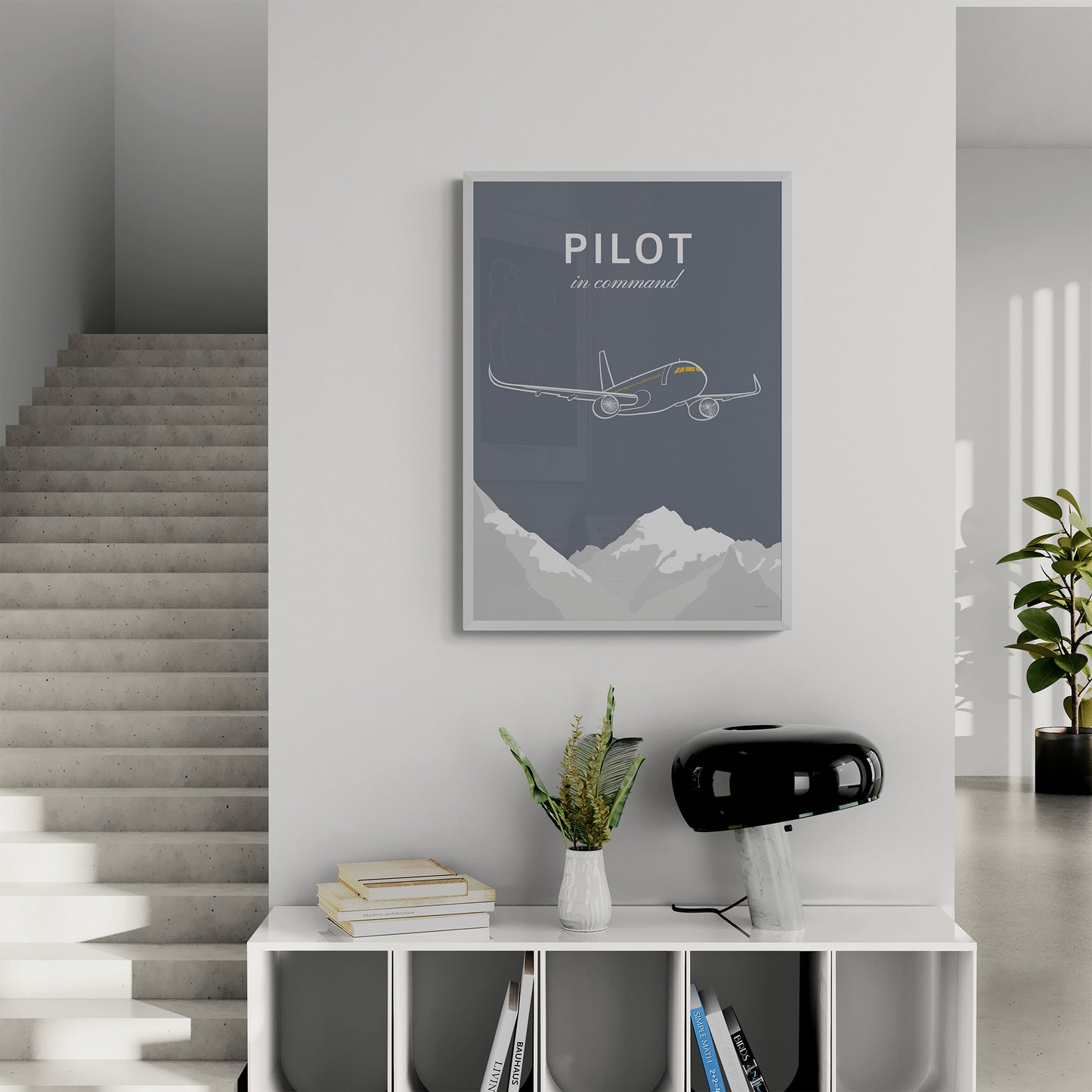 A framed print hanging in a landing of a modern and brightly lit house. It shows a white illustration of a passenger aircraft over mountains on a grey background and the words pilot in command