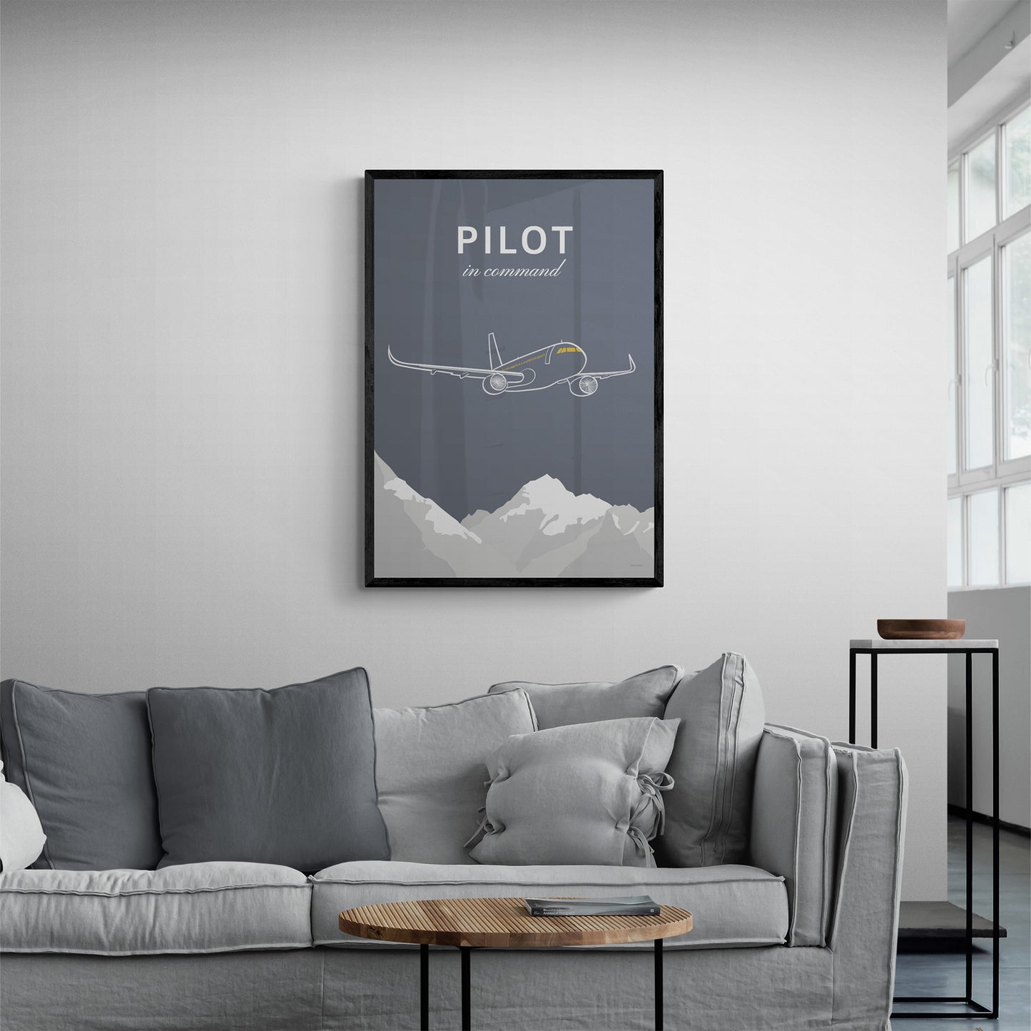 A framed print hanging in a modern looking living room over a light grey sofa with grey cushions. It shows a white illustration of a passenger aircraft over mountains on a grey background and the words pilot in command