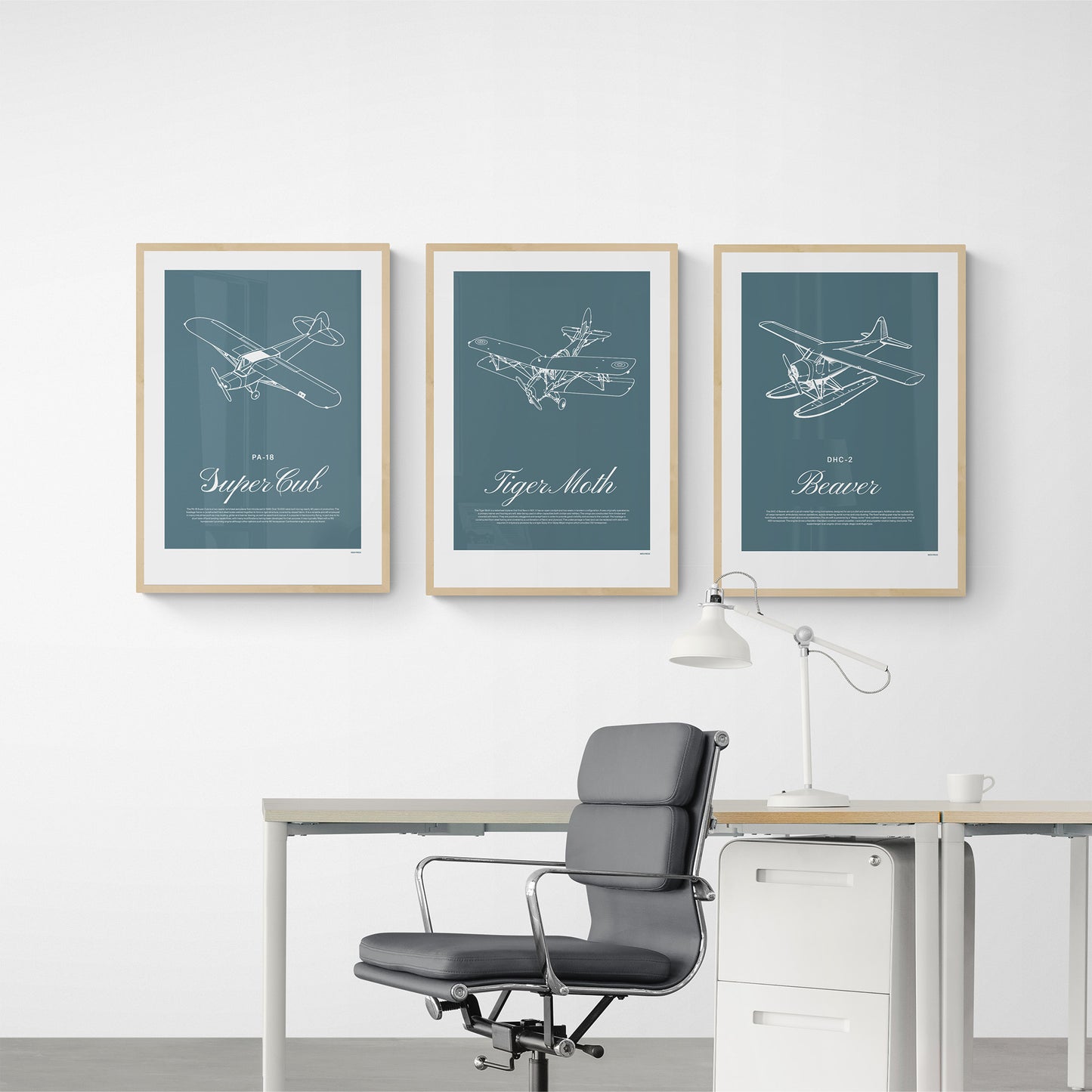 Three framed posters hanging in an office over a desk with a lamp and chair. Each shows a white illustration on a teal background of a light aircraft - DHC-2 Beaver, Tiger Moth, PA-18 Super Cub