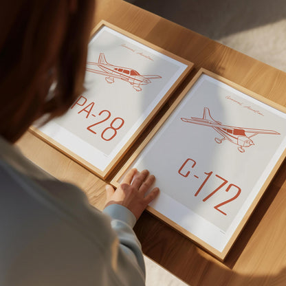 Two framed posters on a table viewed from over the shoulder of a man who has his hand on the corner of one. Each shows a light aircraft illustration with the words General Aviation and the aircraft name in red