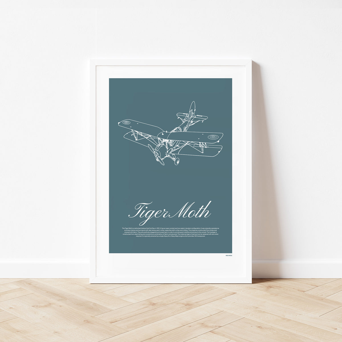 Framed poster standing on wood flooring, leaning against a white wall. It shows a light aircraft illustration with the words Tiger Moth in white over a teal background