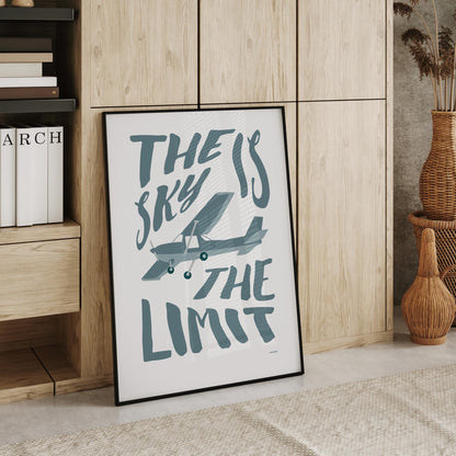 Large framed poster leaning against a cupboard in a home office showing an illustration of a light aircraft with the words THE SKY IS THE LIMIT composed around it in blue