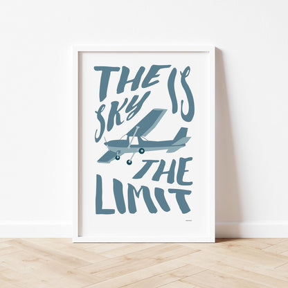 Framed poster standing on wood flooring, leaning against a white wall. It shows an illustration of a light aircraft with the words THE SKY IS THE LIMIT composed around it in blu