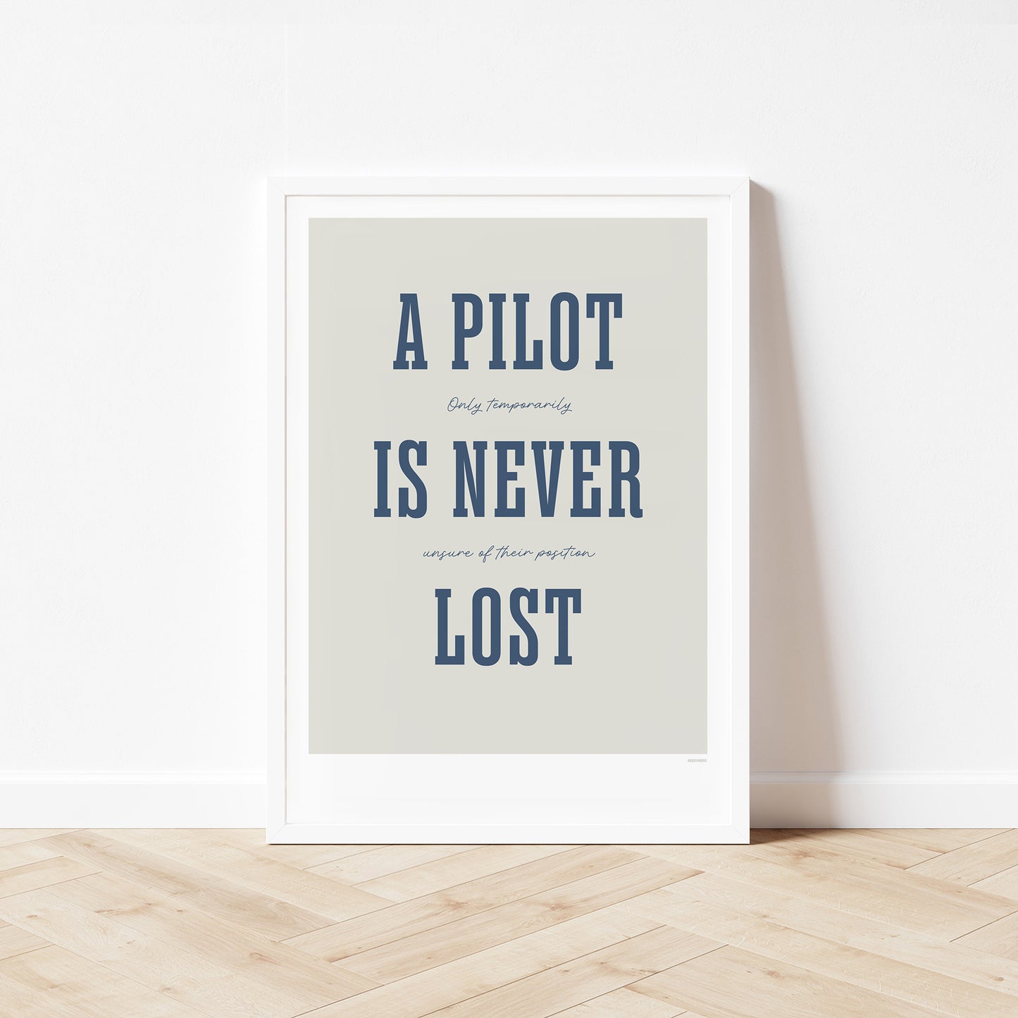 Framed poster standing on wood flooring, leaning against a white wall. It shows the words - A Pilot Is Never Lost - in large blue letters and the words - Only temporarily unsure of their position - in in black handwritten style