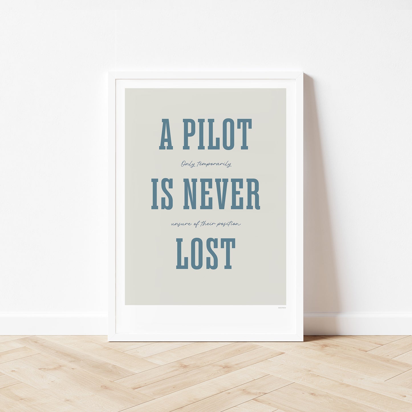 Framed poster standing on wood flooring, leaning against a white wall. It shows the words - A Pilot Is Never Lost - in large blue letters and the words - Only temporarily unsure of their position - in in black handwritten style