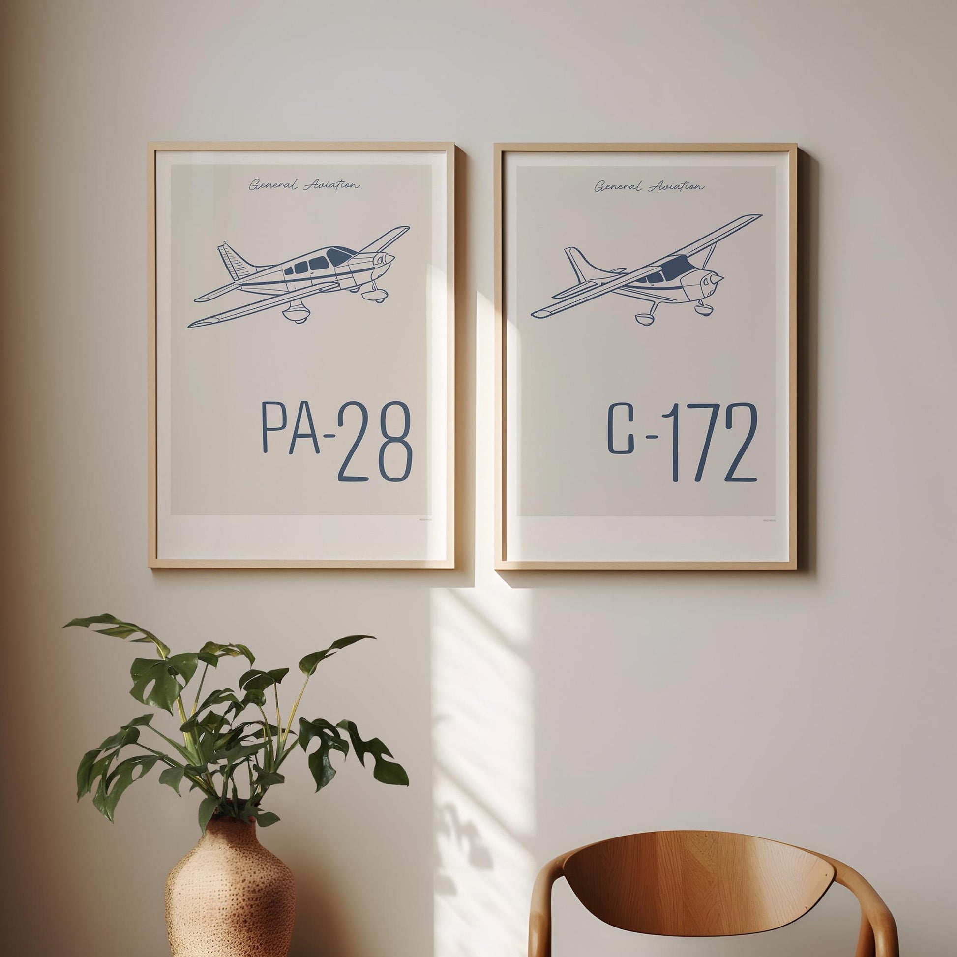 Two framed posters hanging on a white wall above a chair and a plant. Each shows a light aircraft illustration with the words General Aviation and the aircraft name in blue