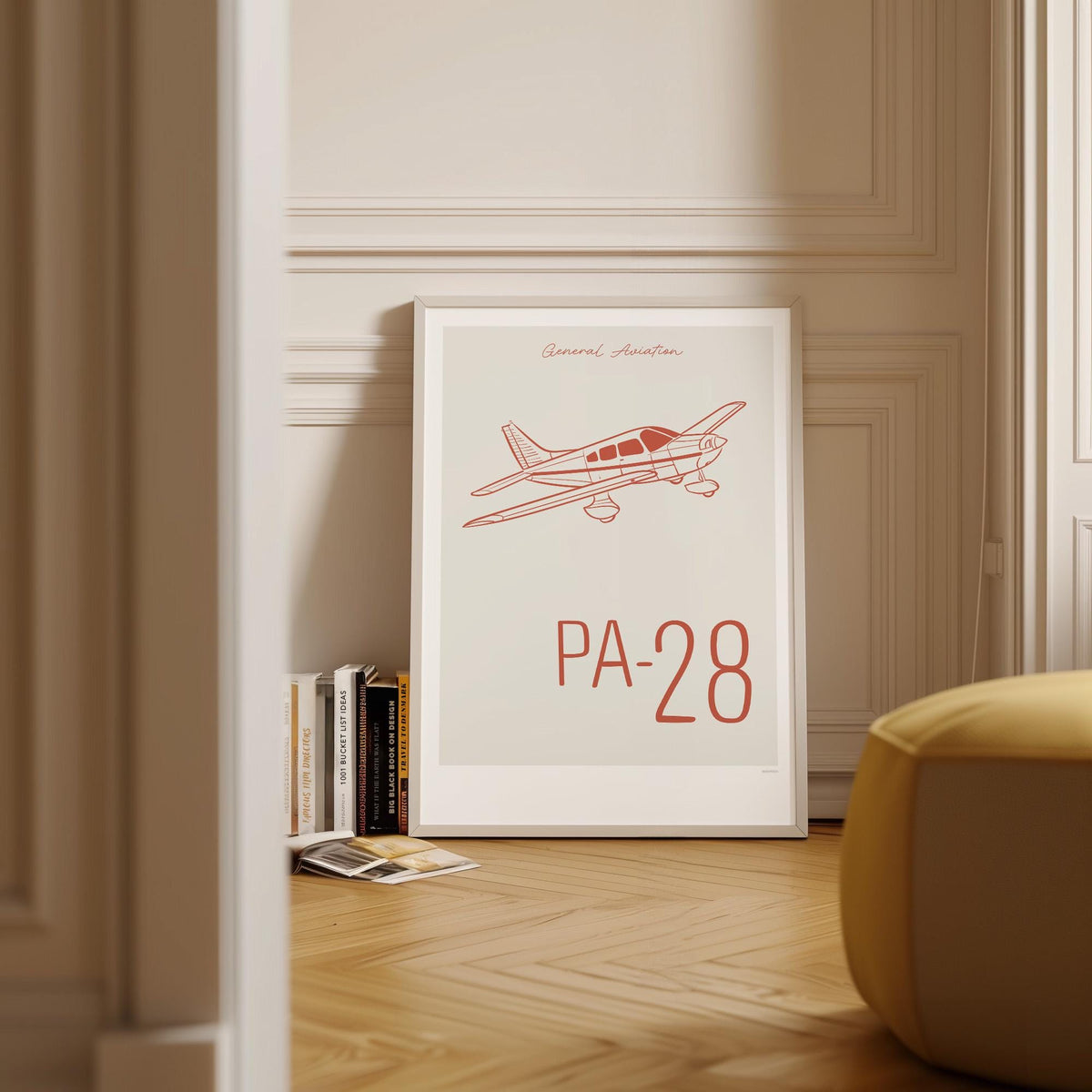 Framed poster on wood flooring leaning against a wall with Scandinavian decor and books around it. It shows a light aircraft illustration with the words General Aviation and PA-28 in red