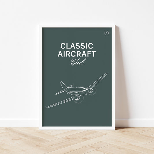 Framed poster standing on wood flooring, leaning against a white wall. It shows a white illustration of a vintage aircraft in a dark green background with the words Classic Aircraft Club