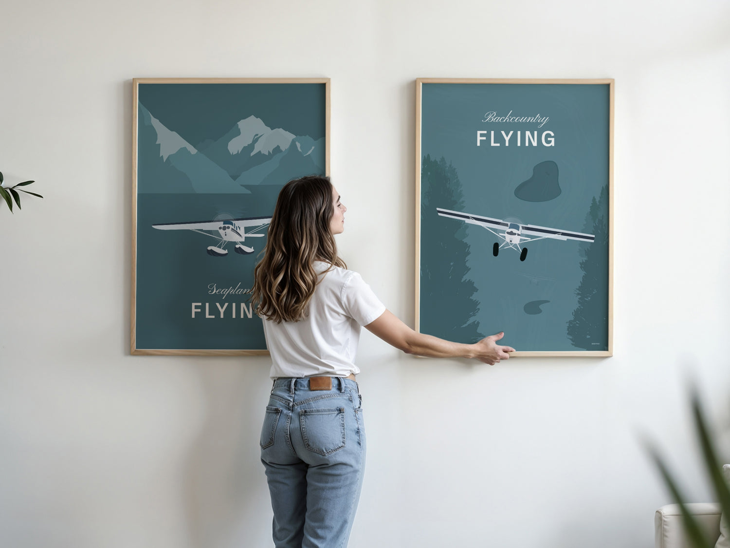 Two large framed posters with a woman who is just finishing to hang one of them in a white wall  in a living room. Each shows a white illustration of a light aircraft with nature elements and a teal coloured background. One has the words backcountry flying and the other seaplane flying 