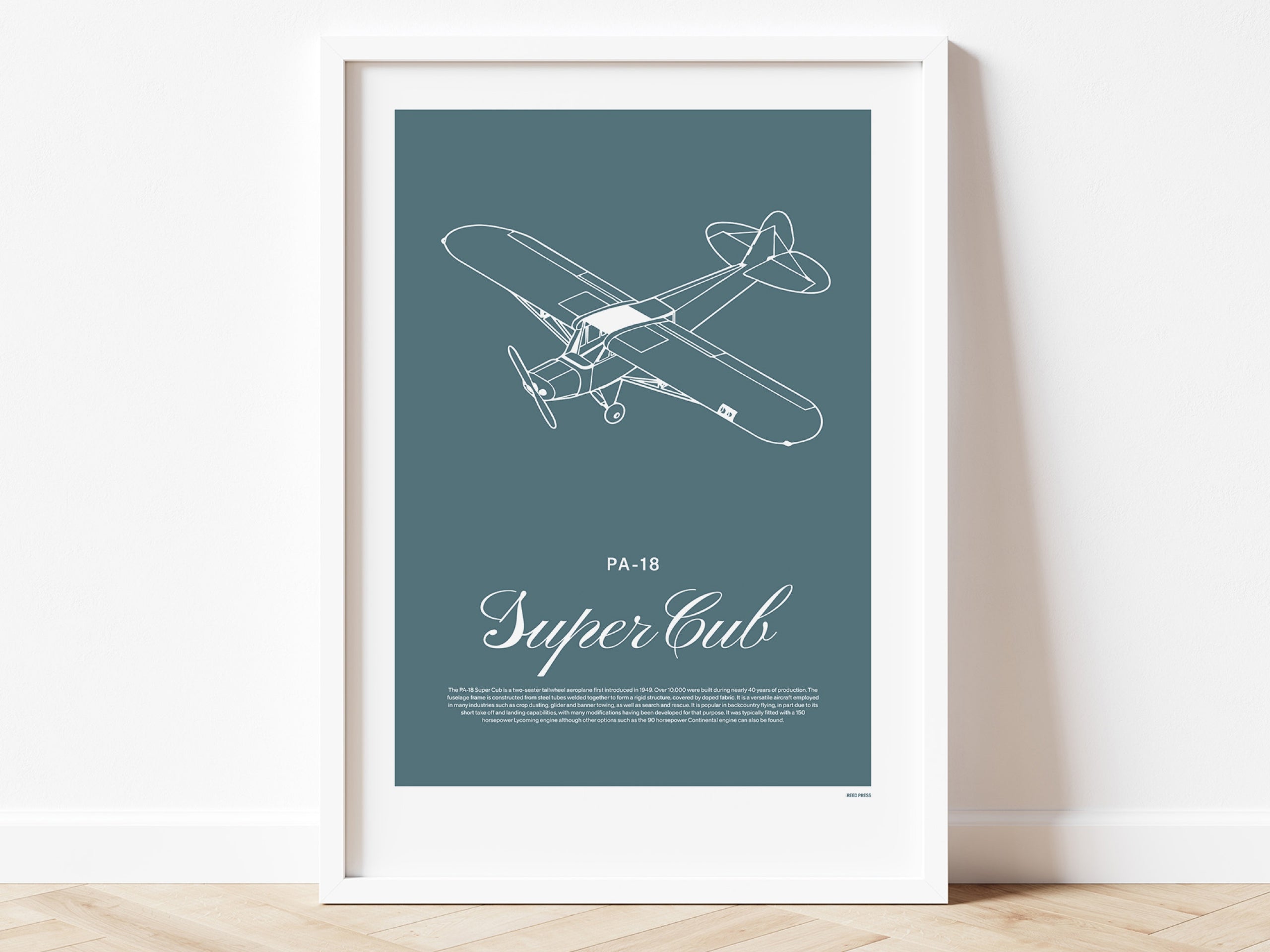 Framed poster standing on wood flooring, leaning against a white wall. It shows a light aircraft illustration with the words PA-18 Super Cub in white over a teal background