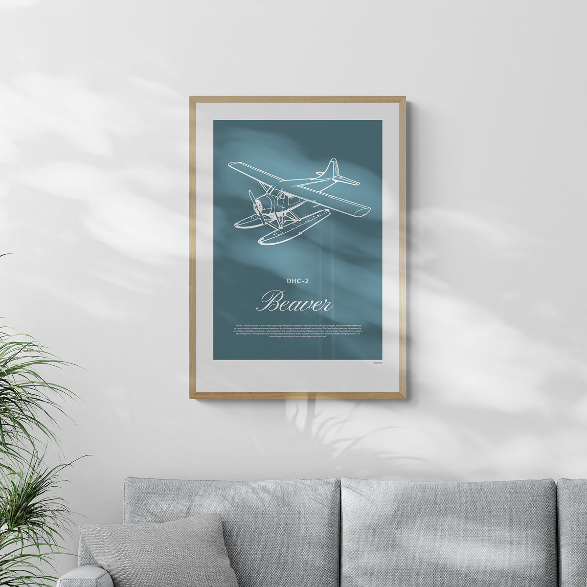 A framed print hanging in a living room, over a grey sofa. It shows a light aircraft illustration with the words DHC-2 Beaver in white over a teal background