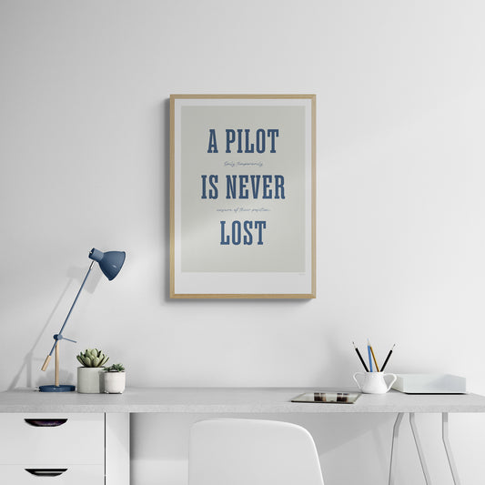 A framed print hanging in a home office over a white desk and chair. It shows the words - A Pilot Is Never Lost - in large blue letters and the words - Only temporarily unsure of their position - in in black handwritten style