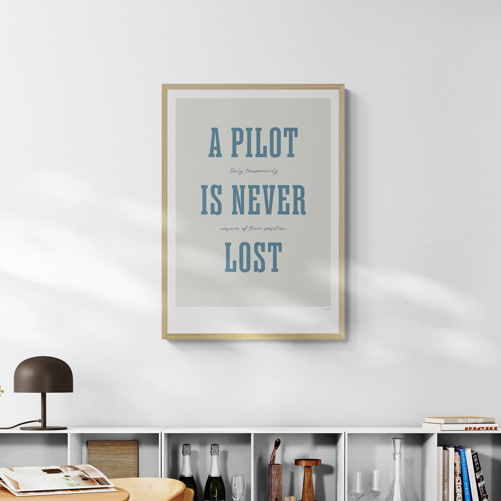 A framed print hanging over a bookshelf. It shows the words - A Pilot Is Never Lost - in large blue letters and the words - Only temporarily unsure of their position - in in black handwritten style