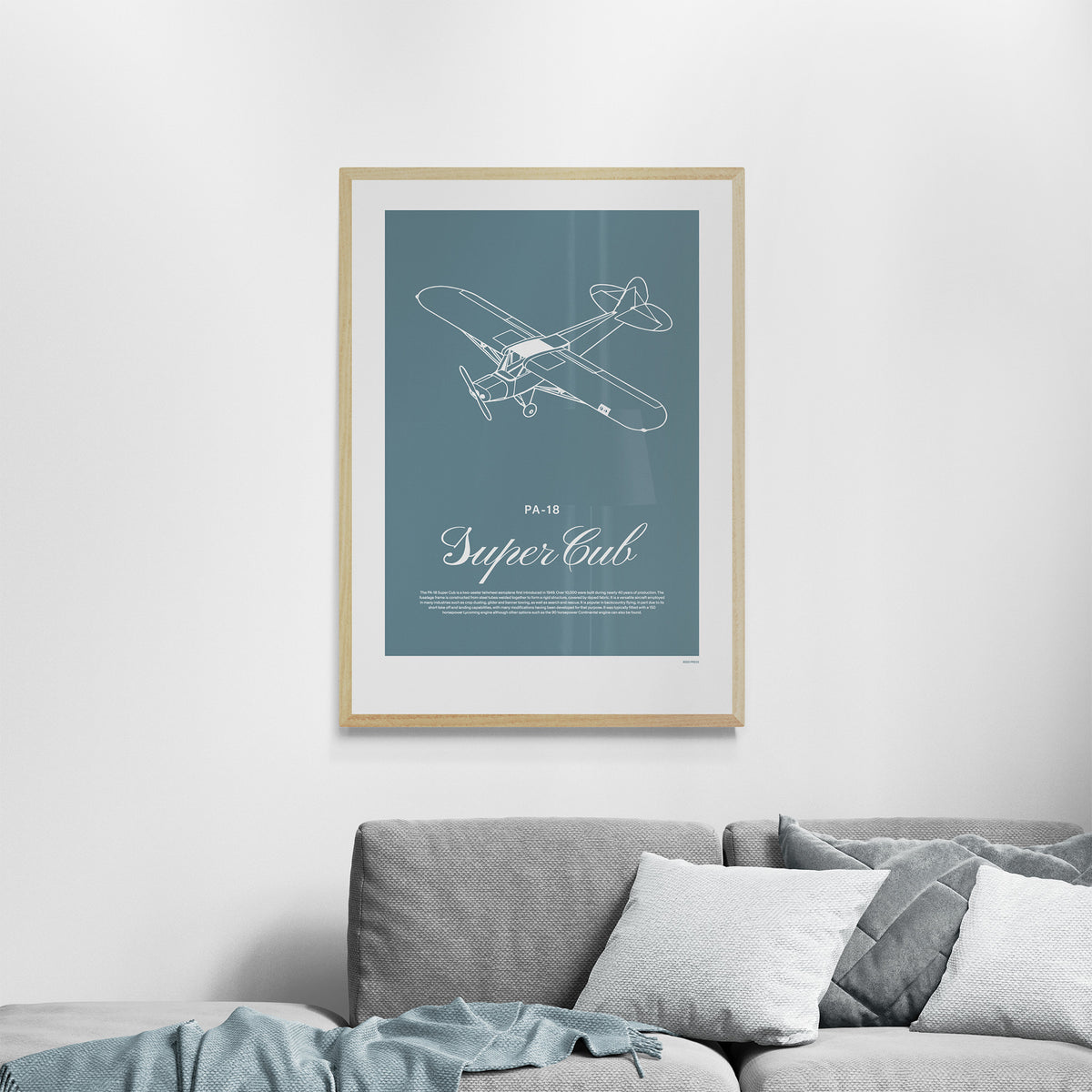A framed print hanging over a grey sofa. It shows a light aircraft illustration with the words PA-18 Super Cub in white over a teal background