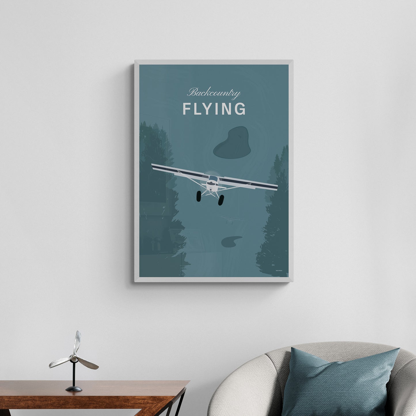 A framed print hanging over a chair and a console table with a propeller shaped ornament. It shows a white illustration of a light aircraft with trees around it and a a teal coloured background and the words backcountry flying