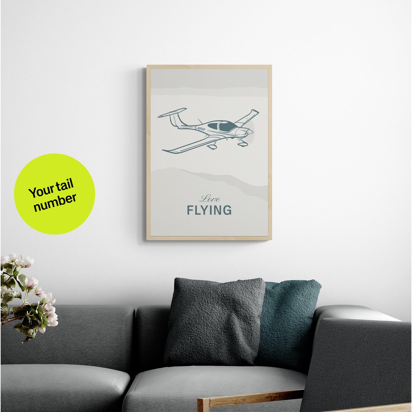 A framed print hanging over a grey sofa with cushions. It shows a light aircraft illustration with the words Love Flying in white over an off white background