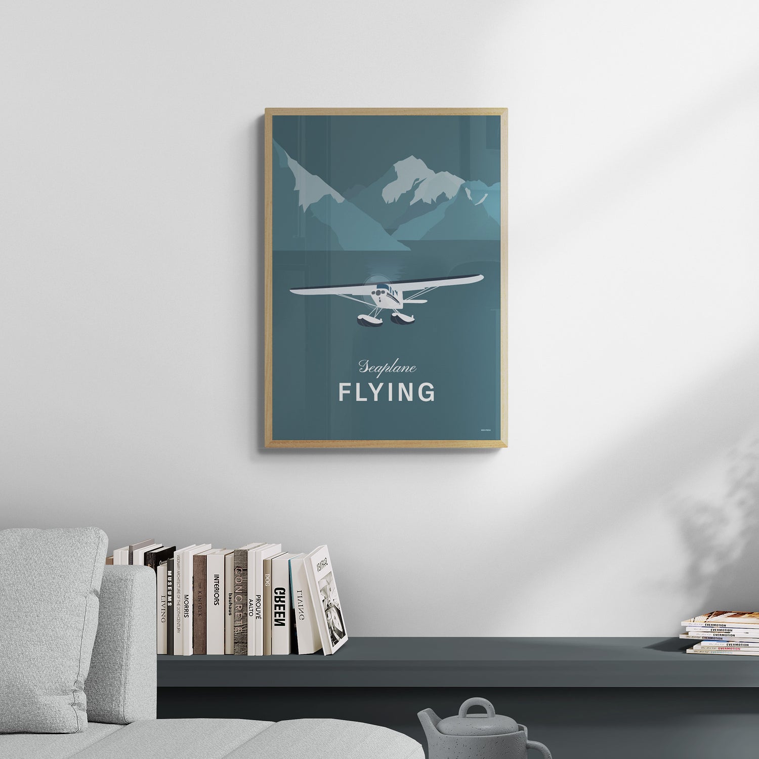 A framed print hanging in a living room over a sofa and bookshelf. It shows a white illustration of a seaplane with mountains in a teal coloured background and the words seaplane flying