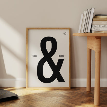 A framed poster leaning against a white wall on a wood floor next to a table with books. It shows the words stick & rudder in black with the & being large and stick/rudder being small 