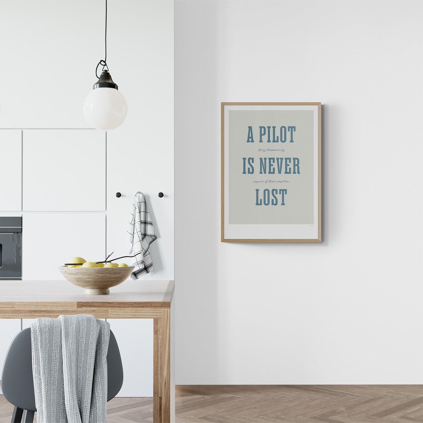 Framed print hanging in a modern kitchen. It shows the words - A Pilot Is Never Lost - in large blue letters and the words - Only temporarily unsure of their position - in in black handwritten style