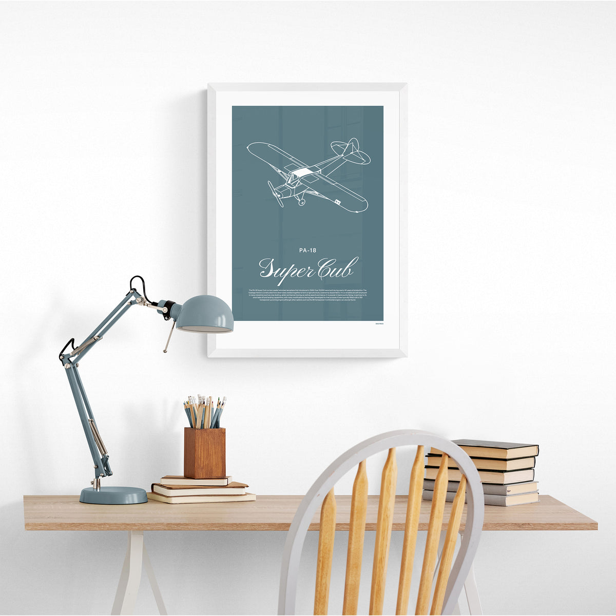 A framed print hanging in a home office over a desk and chair with a lamp. It shows a light aircraft illustration with the words PA-18 Super Cub in white over a teal background