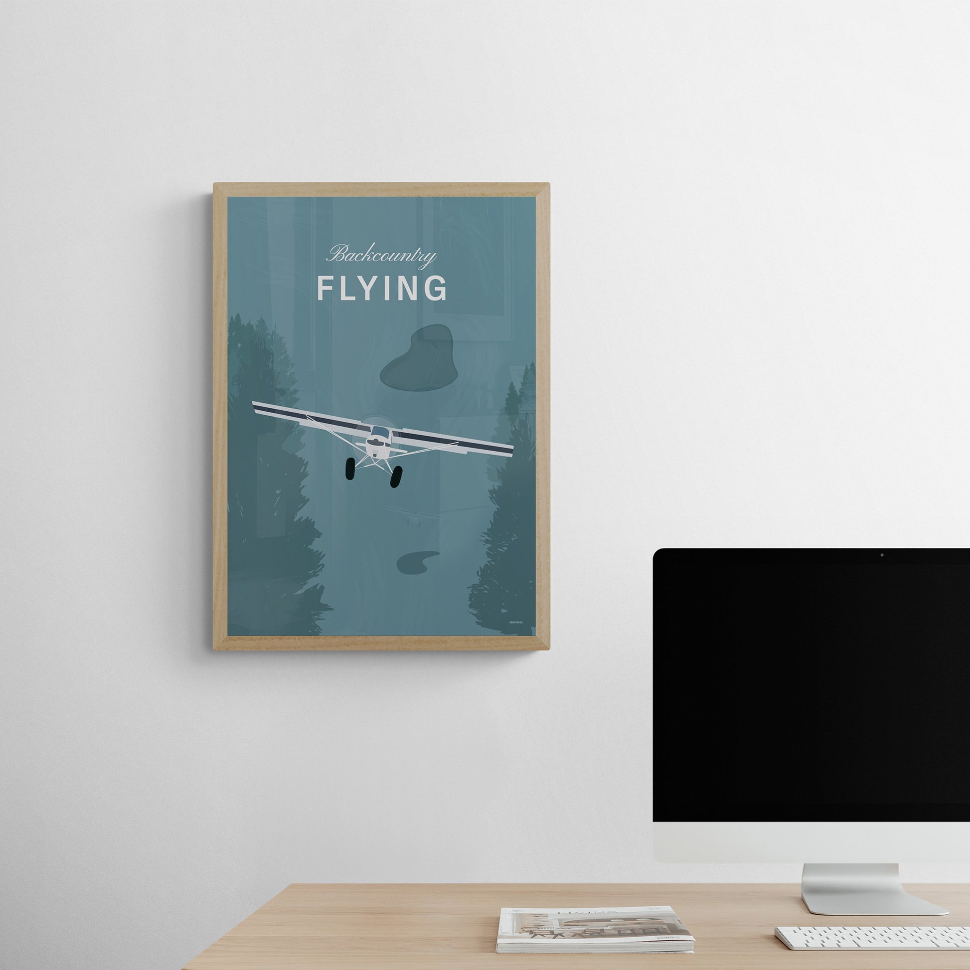 A framed print hanging on a study over a desk with a computer. It shows a white illustration of a light aircraft with trees around it and a a teal coloured background and the words backcountry flying