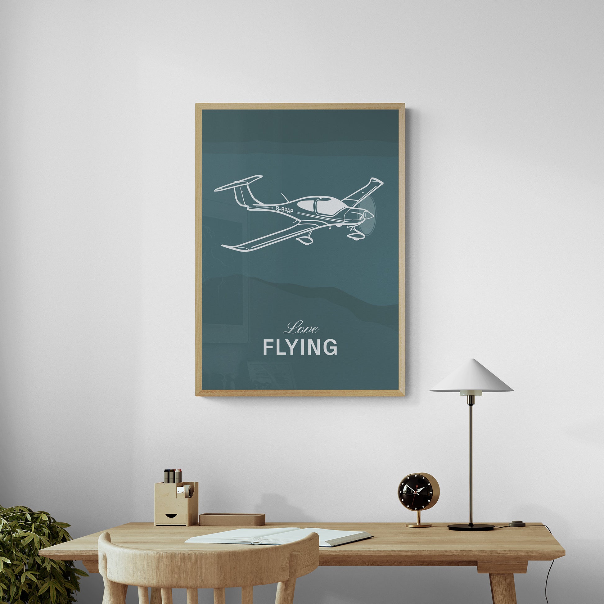 A framed print hanging over a desk and chair in a home office. It shows a light aircraft illustration with the words Love Flying in white over a teal background
