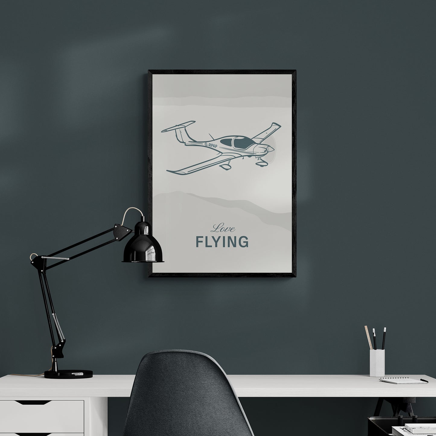 A framed print hanging over a desk and a chair, next to a lamp. It shows a light aircraft illustration with the words Love Flying in white over an off white background