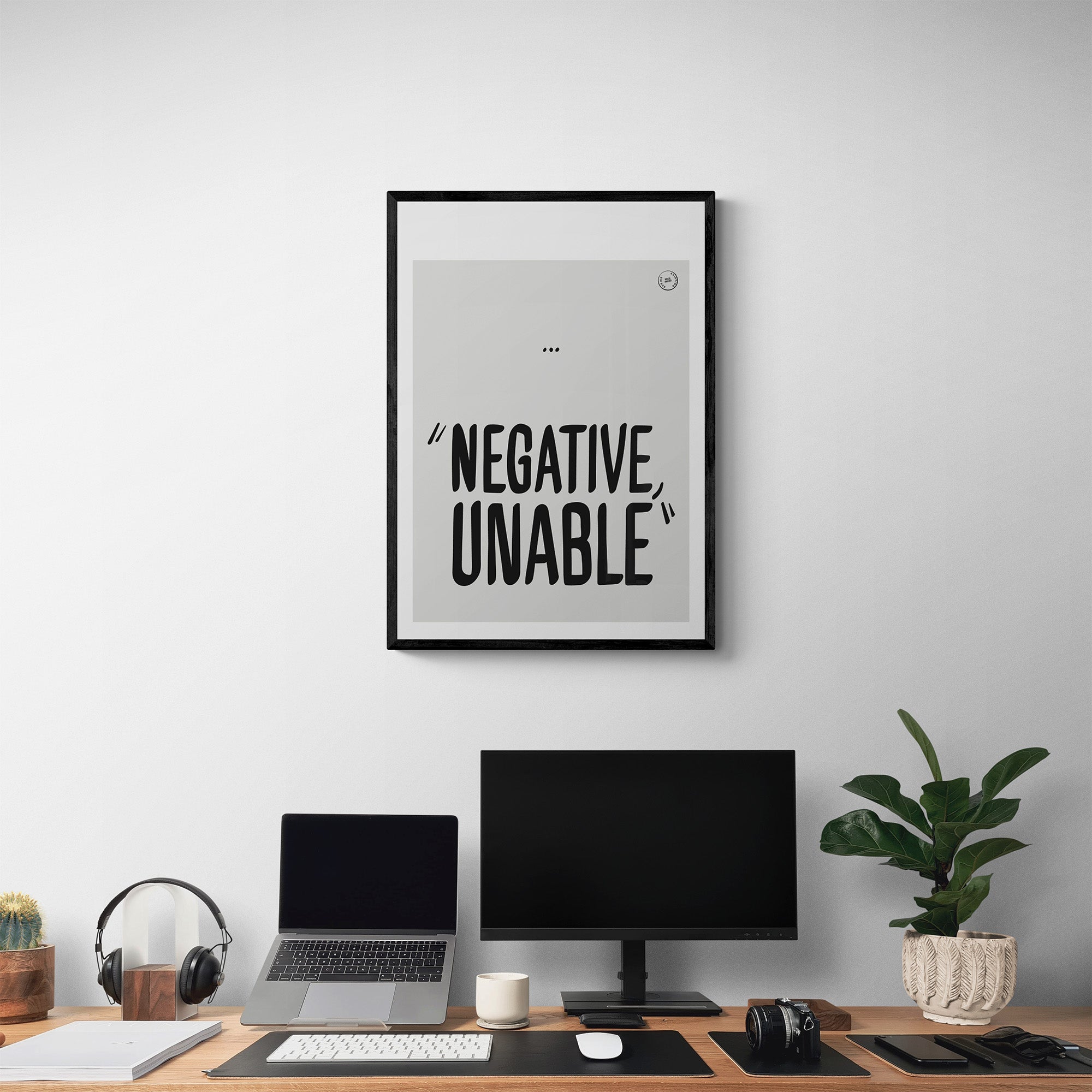 A framed poster hanging in a home office over a desk set up with computers. It shows the words negative, unable in black hand drawn letters 