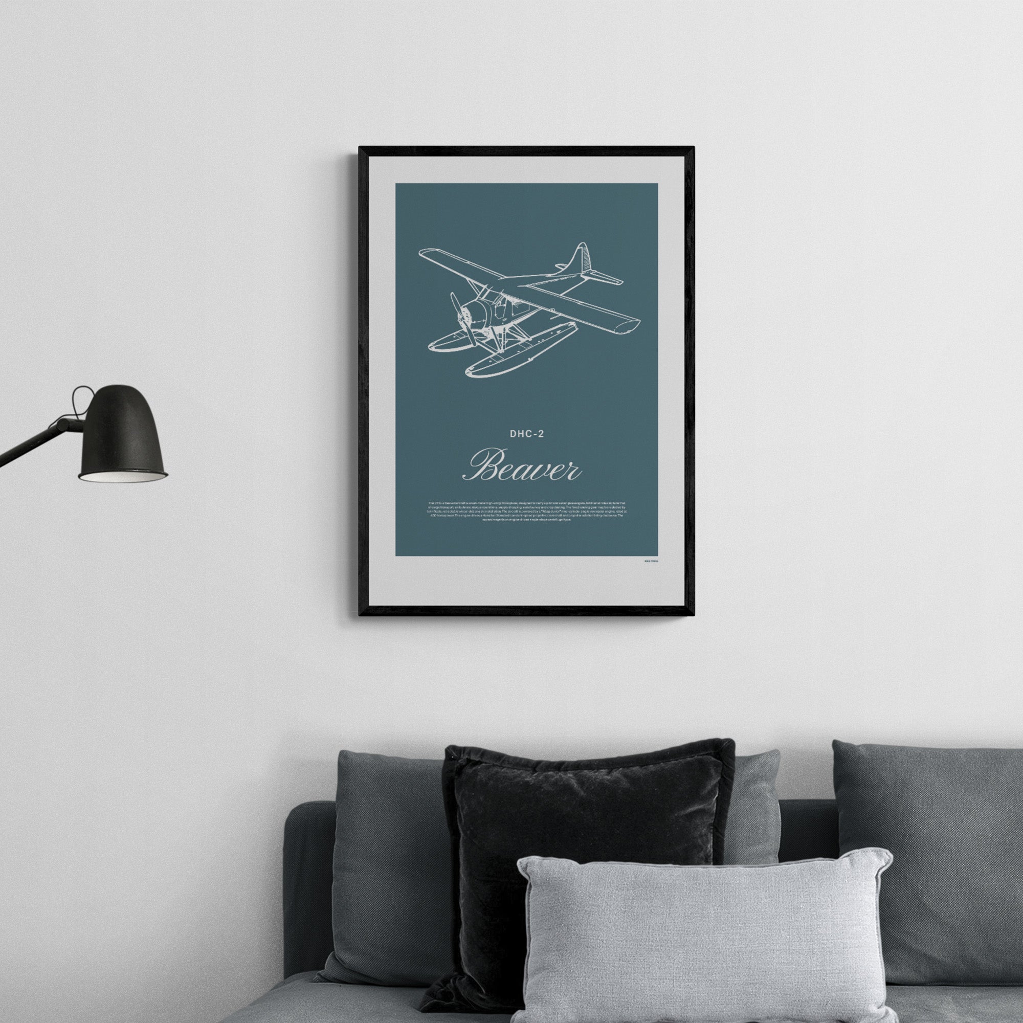 A framed print hanging over a grey sofa. I shows a light aircraft illustration with the words DHC-2 Beaver in white over a teal background