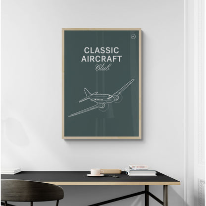 A framed poster hanging on a home office over a desk and chair. It shows a white illustration of a vintage aircraft in a dark green background with the words Classic Aircraft Club