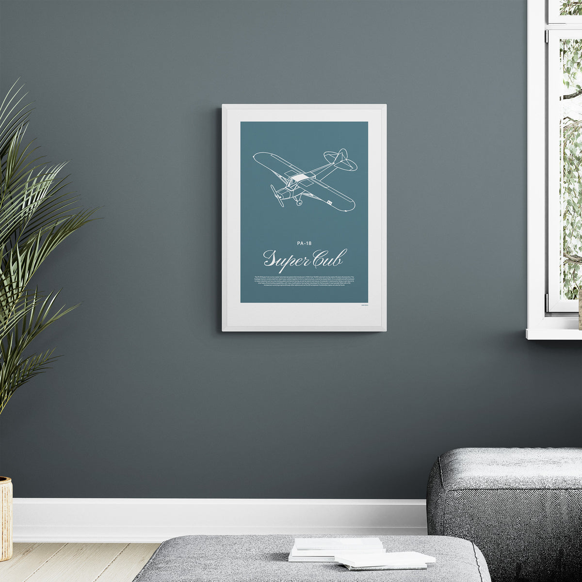 A framed print hanging on a dark grey wall in a modern living room. It shows a light aircraft illustration with the words PA-18 Super Cub in white over a teal background