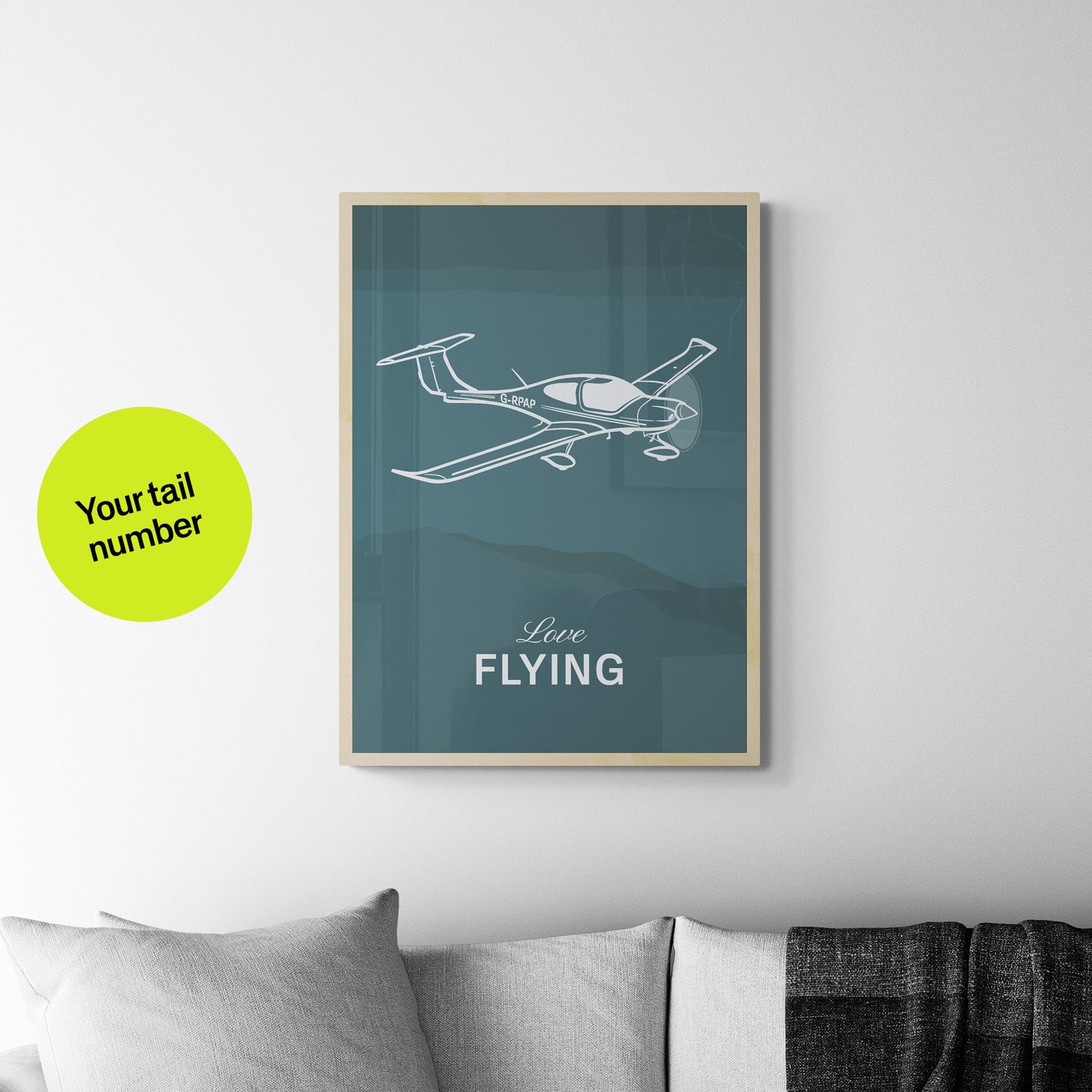 A framed print hanging over a light grey sofa. It shows a light aircraft illustration with the words Love Flying in white over a teal background