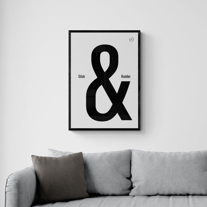 A framed print hanging over a grey sofa. It shows the words stick & rudder in black with the & being large and stick/rudder being small 