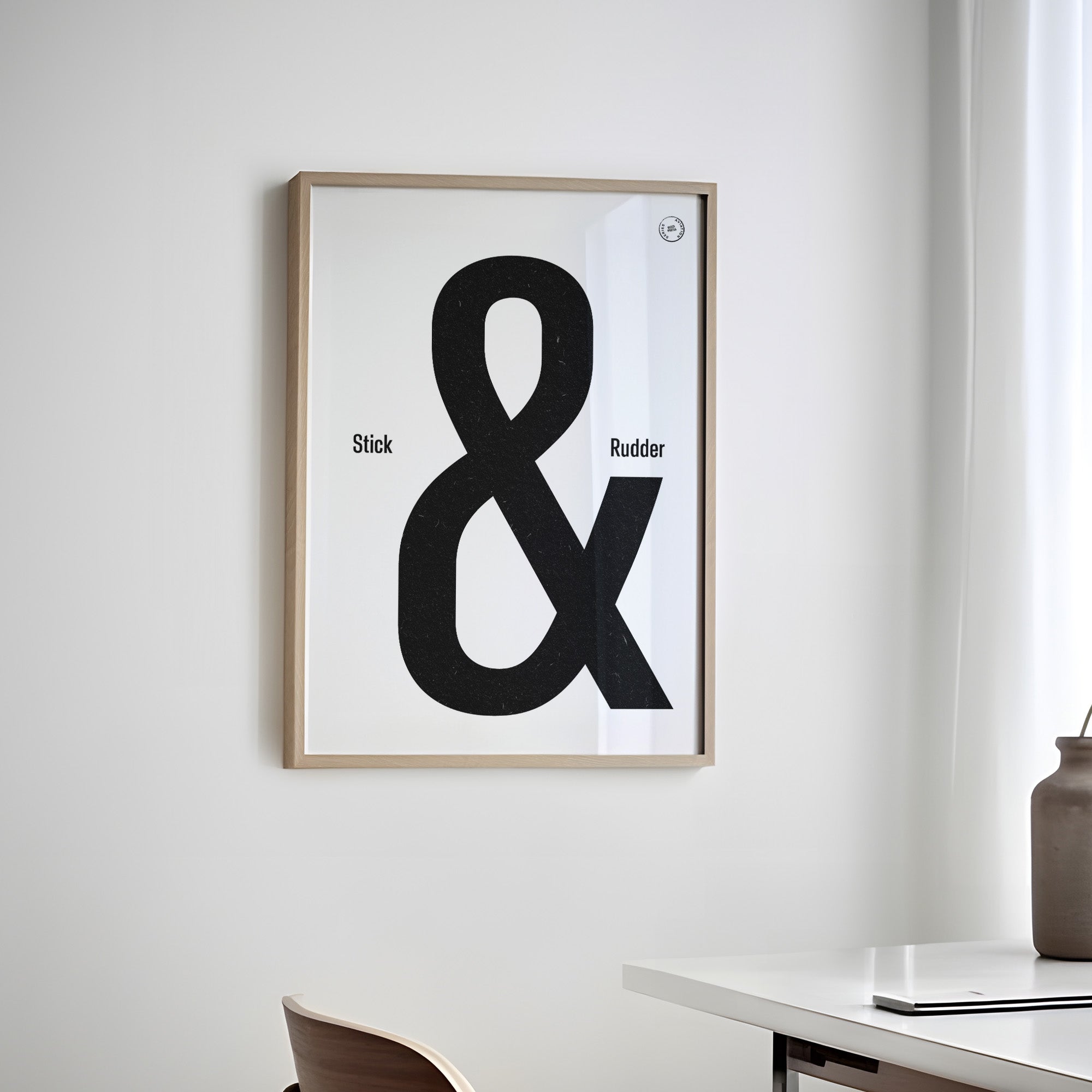 Framed poster hanging on a white wall of a home office with a table and chair. It shows the words stick & rudder in black with the & being large and stick/rudder being small 