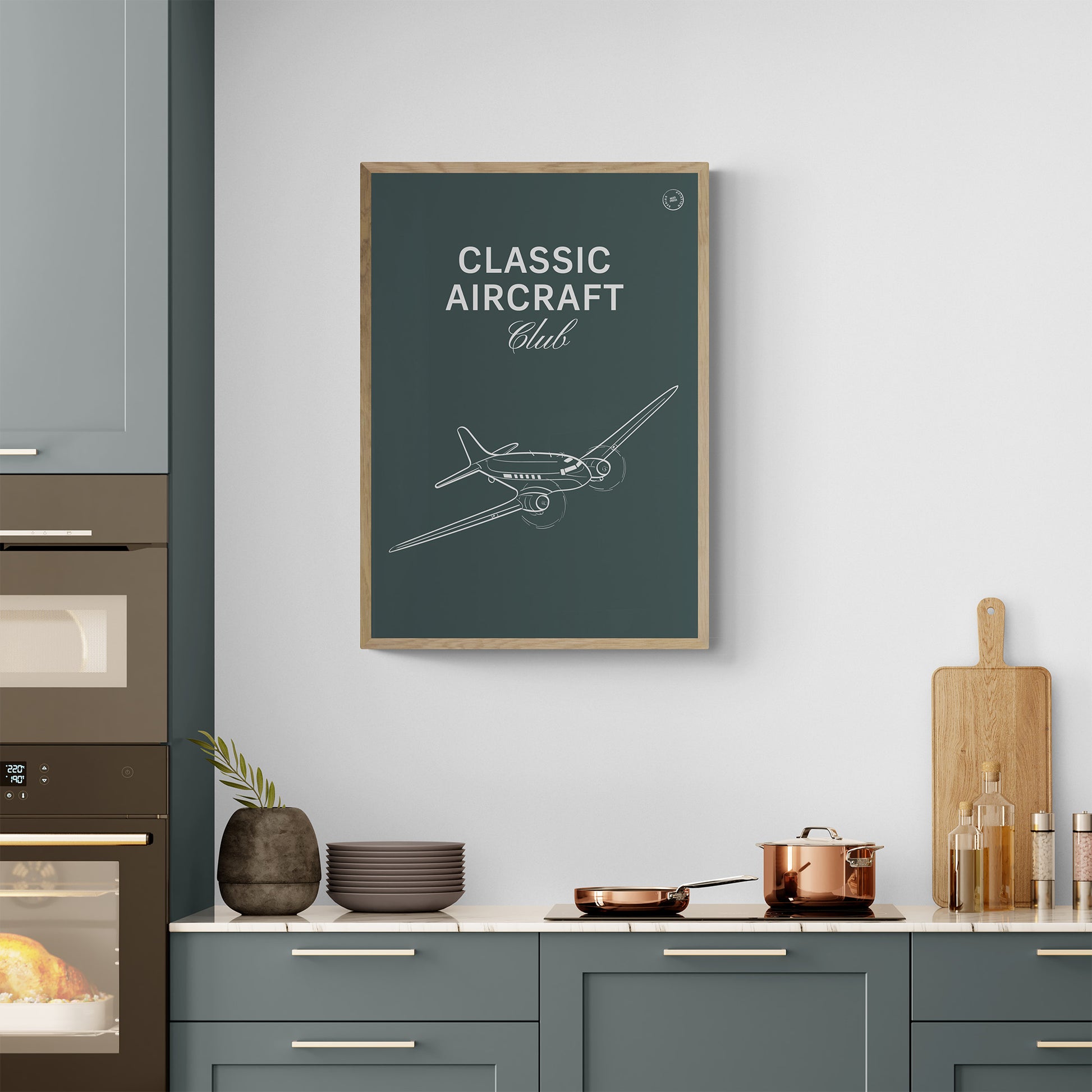 A framed poster hanging on a modern kitchen. It shows a white illustration of a vintage aircraft in a dark green background with the words Classic Aircraft Club