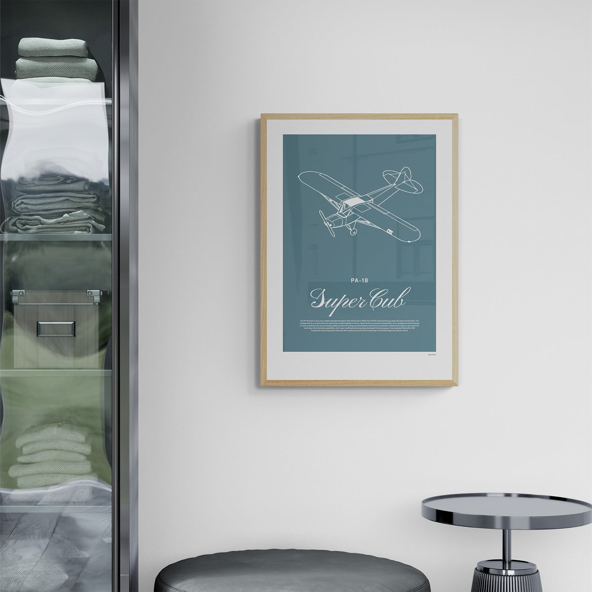 A framed print hanging next to a wardrobe over a side table and a stool. It shows a light aircraft illustration with the words PA-18 Super Cub in white over a teal background