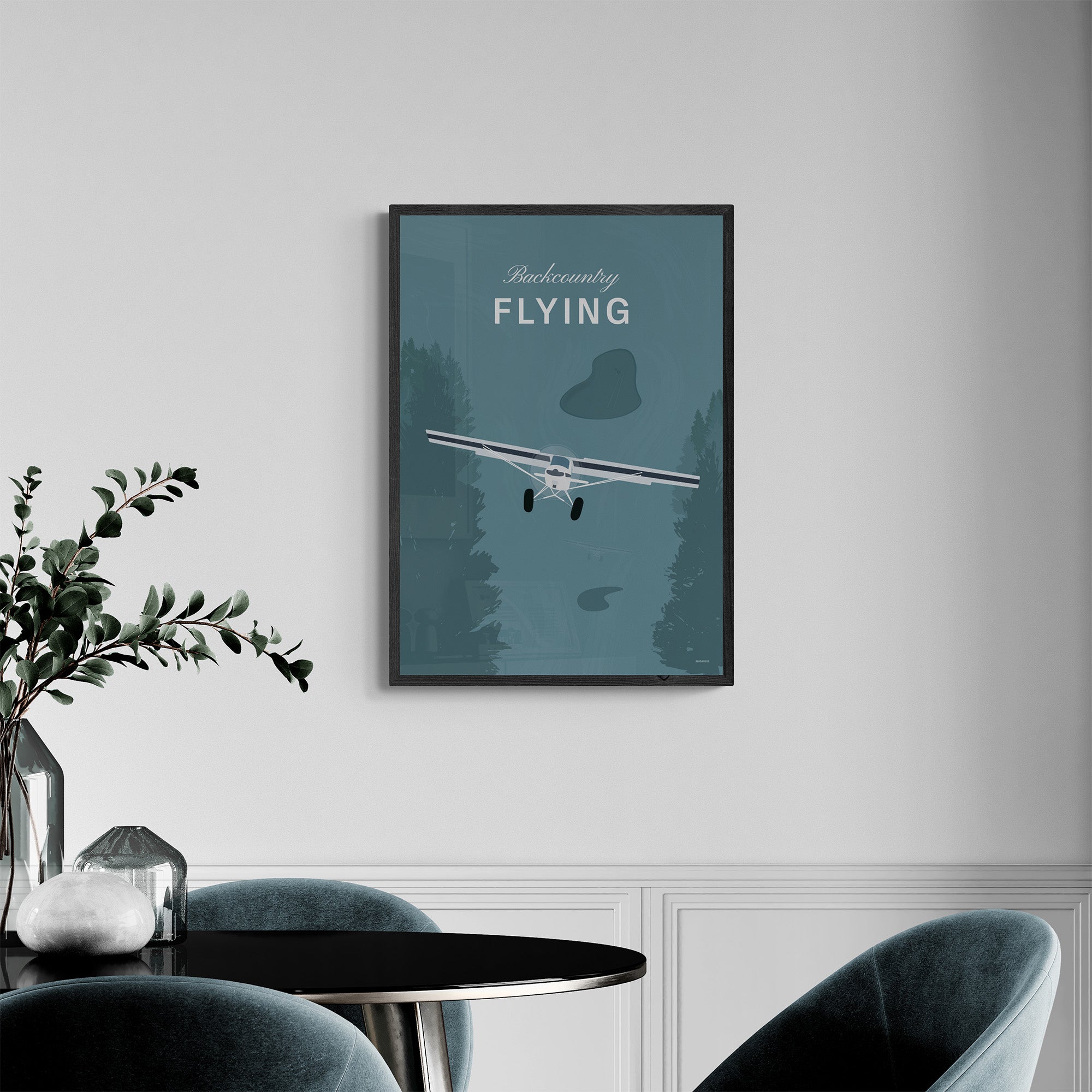 A framed print hanging on a dining room over a circular table and chairs. It shows a white illustration of a light aircraft with trees around it and a a teal coloured background and the words backcountry flying