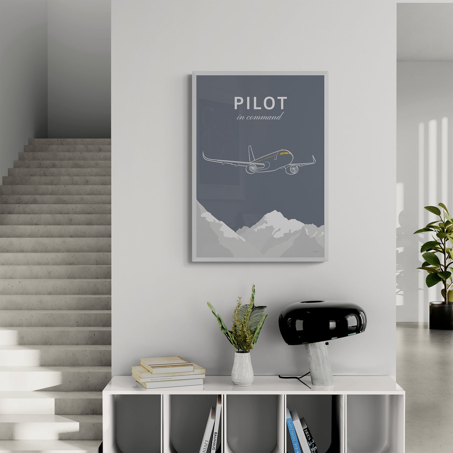 A framed print hanging in a landing of a modern and brightly lit house. It shows a white illustration of a passenger aircraft over mountains on a grey background and the words pilot in command 