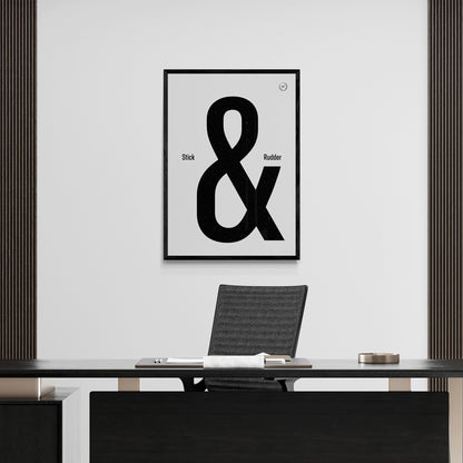 A framed print hanging in a modern office over a desk and chair. It shows the words stick & rudder in black with the & being large and stick/rudder being small 