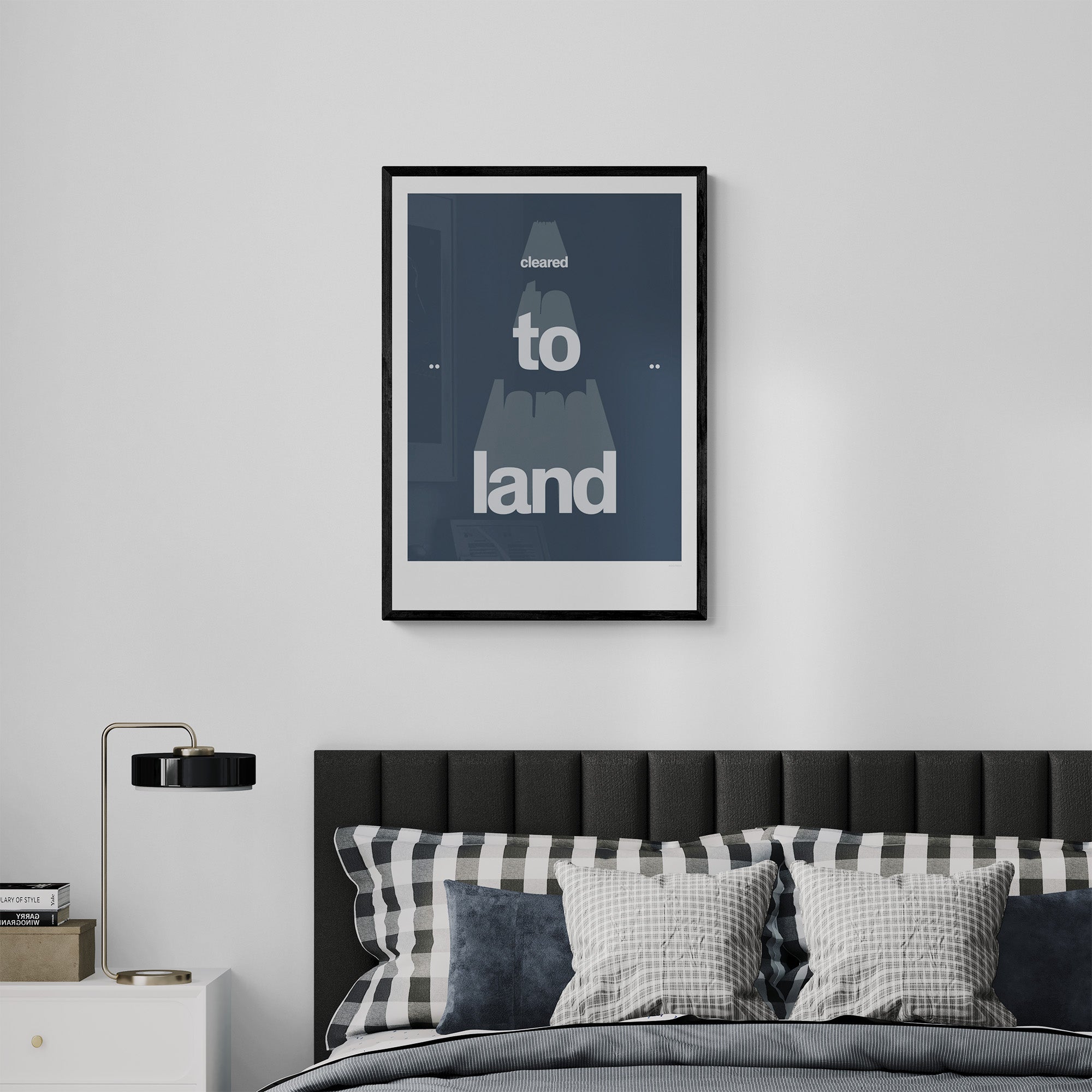 Framed print hanging on a modern bedroom over a bed with pillows and cushions. It shows the words cleared to land in white with a dark blue background 