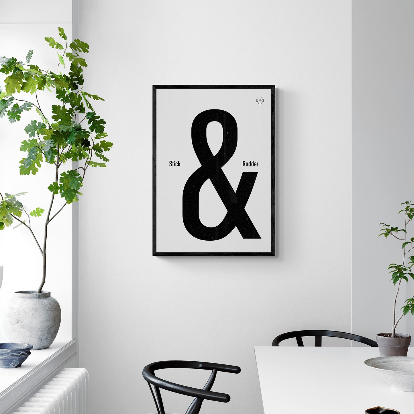 A framed print hanging in a modern bright dining room over a desk and chairs. It shows the words stick & rudder in black with the & being large and stick/rudder being small 