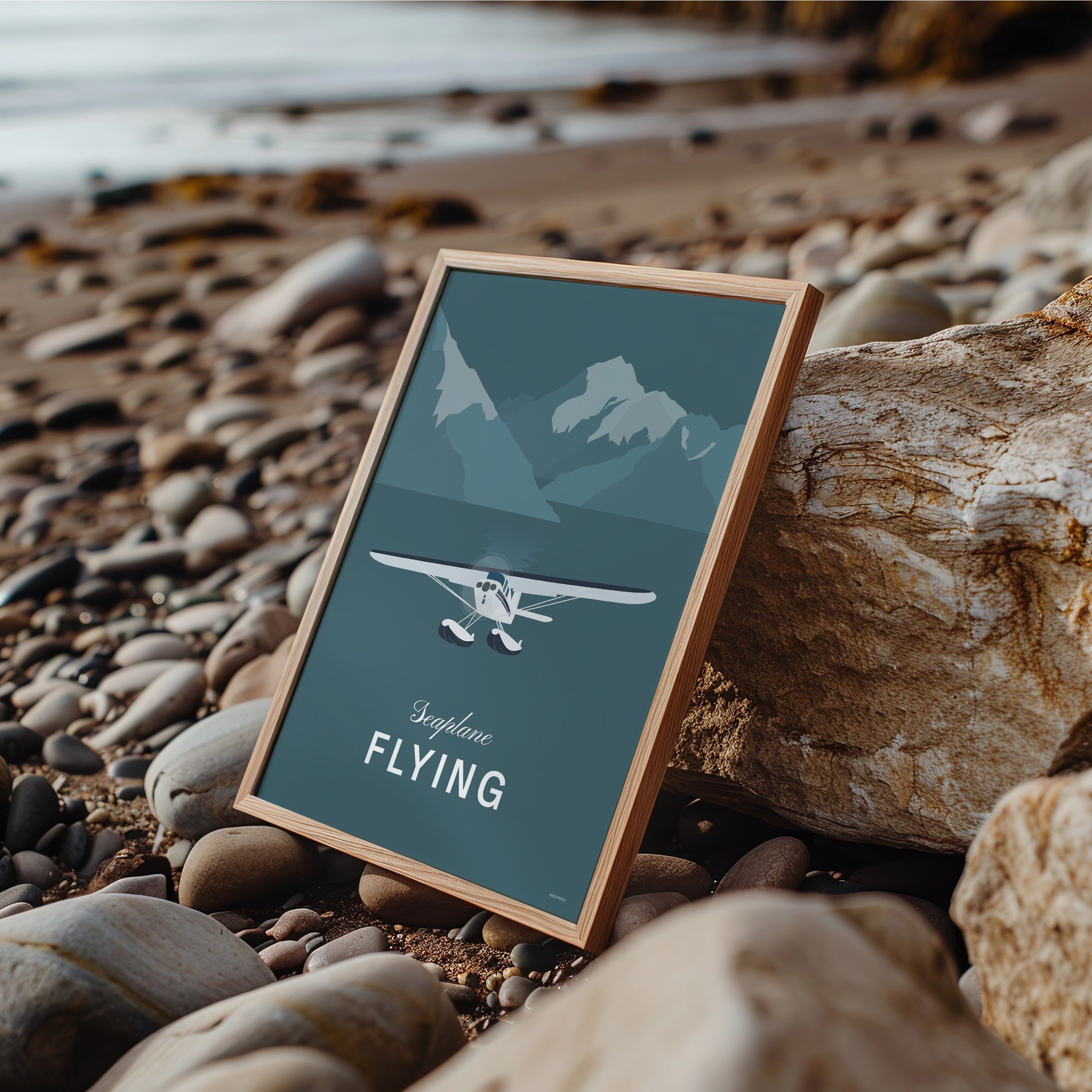 Framed poster leaning against a rock on a shingle beach showing a white illustration of a seaplane with mountains in a teal coloured background and the words seaplane flying